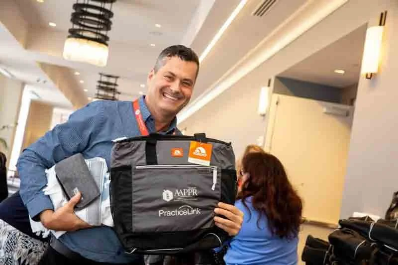 A smiling man holding a black bag with the APPRE PracticeLink logo at a conference or seminar in a well-lit room.