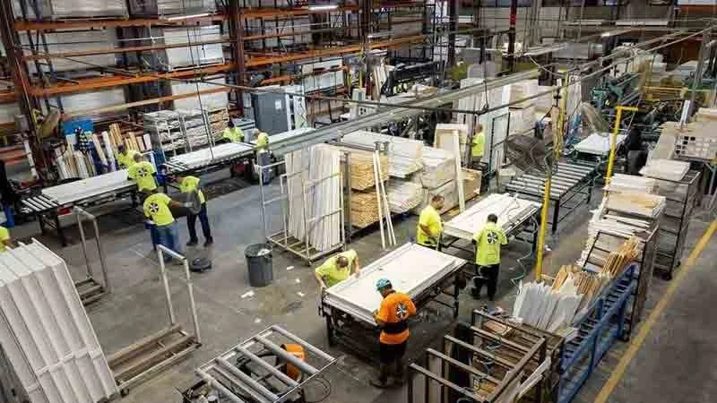 Factory workers in yellow and orange safety shirts operate machinery and handle materials in a large industrial warehouse with steel shelving and a cluttered workspace.