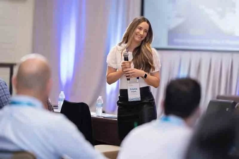 A young woman speaks into a microphone during a conference presentation, smiling, with a badge hanging around her neck, audience members blurred in the foreground.