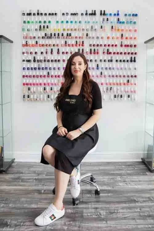A woman sitting on a swivel chair in front of a wall display of numerous nail polish bottles organized by color. She is wearing a black dress and white sneakers with a logo, and smiling at the camera.
