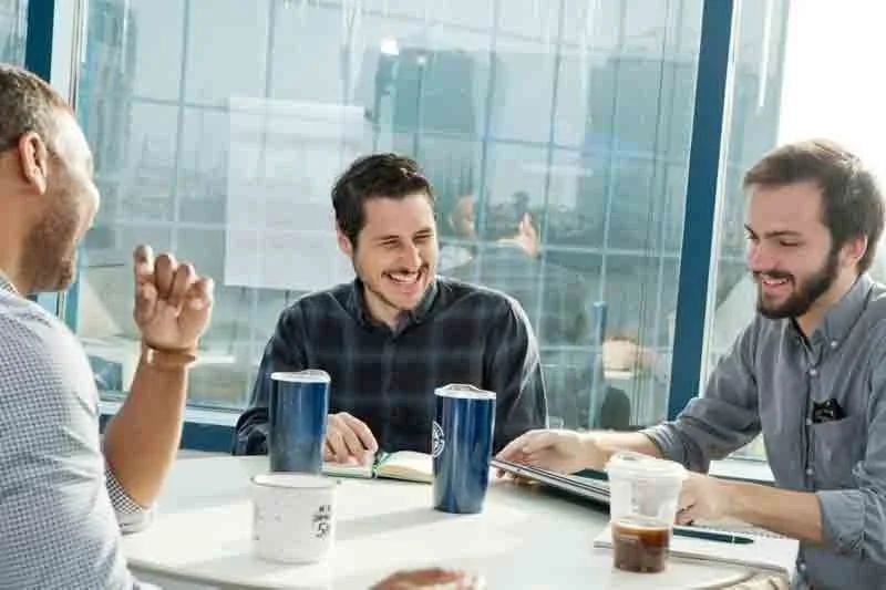 Three men sitting at a table in a modern office, laughing and talking, with cups of coffee or tea, and a glass wall in the background.