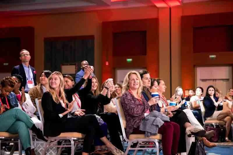Audience members sitting and smiling at a conference or seminar, with some taking photos and others enjoying the presentation.