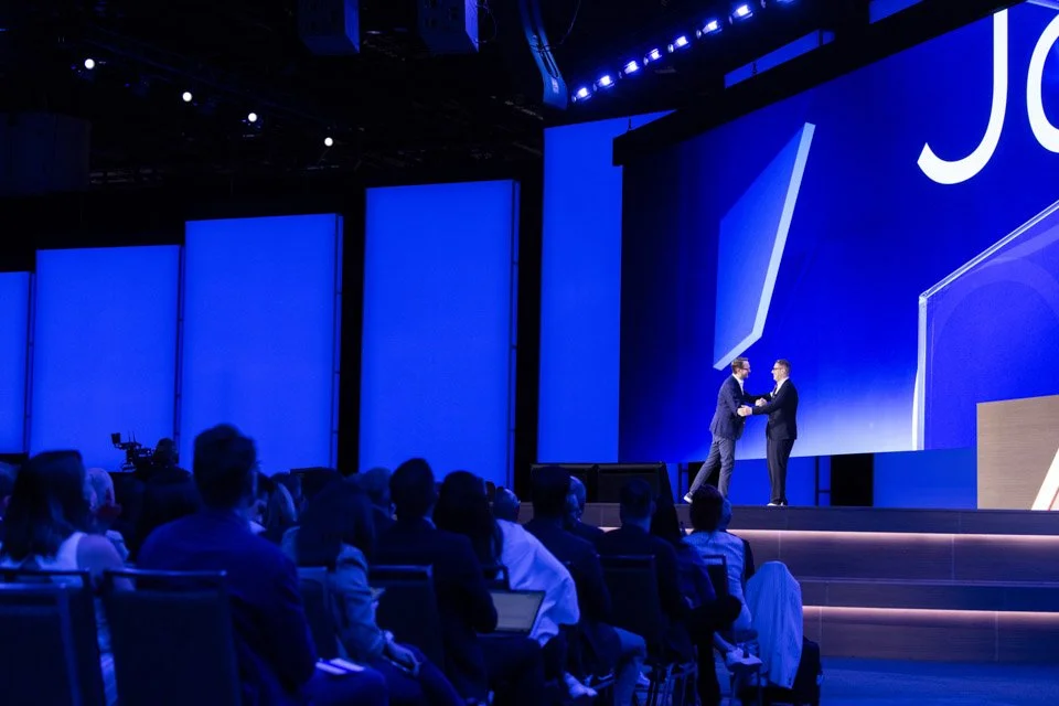 Two men shaking hands on stage at a conference, with a large blue screen behind them and an audience seated in front.