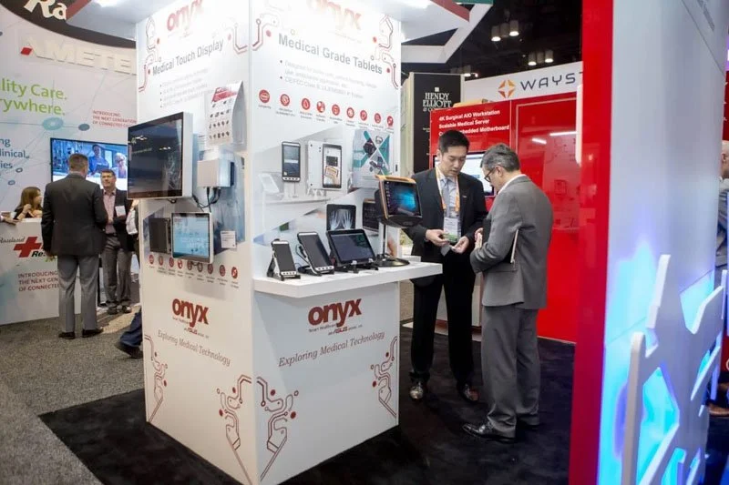 Trade show booth displaying medical tablets and touch screen devices with attendees inspecting products.