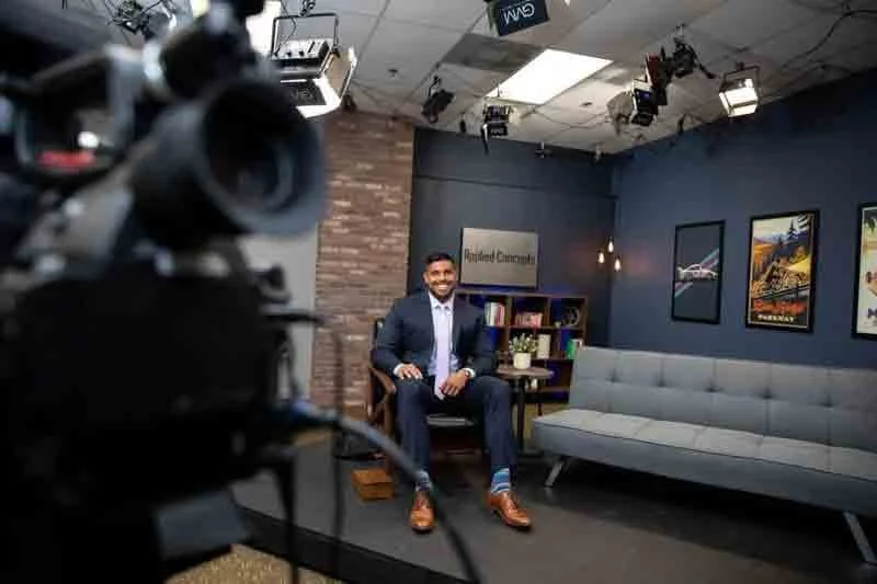 Man in a suit sitting on a chair in a TV studio or office set, with a modern gray sofa and framed pictures on the wall behind him.