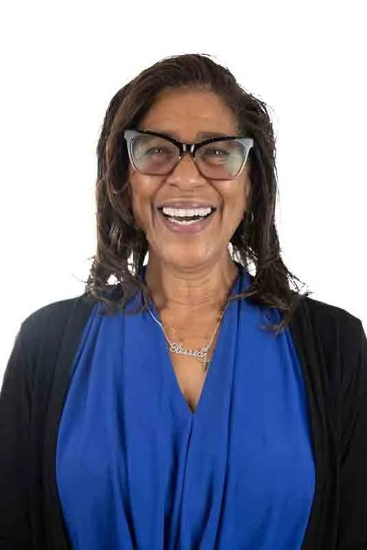 A woman with dark brown hair, glasses, and a bright smile wearing a blue top and a black cardigan.