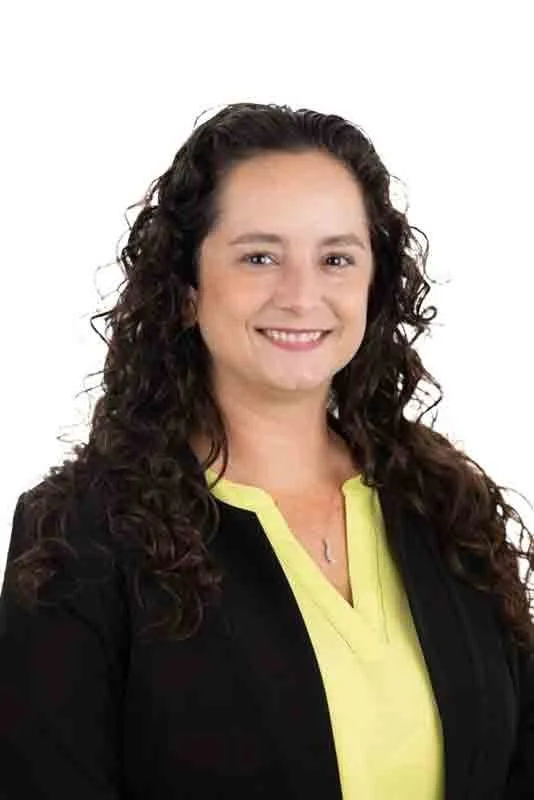A woman with long, curly brown hair smiling, wearing a black blazer over a yellow shirt, against a plain white background.