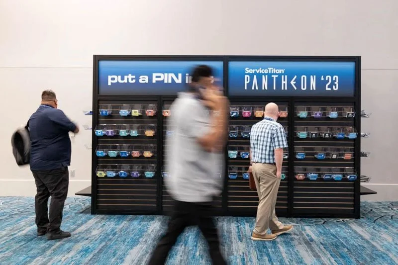 People walking past a booth at ServiceTitan's PANTHEON '23 conference, where a display features various connected devices or cards with a sign that says 'put a PIN in'.