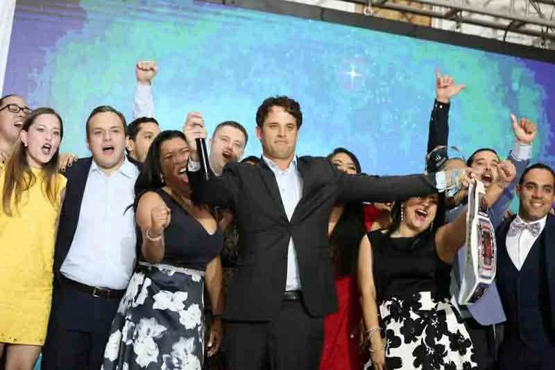 Group of people celebrating on stage, some raising fists, with a man in the center holding a trophy and a boxed prize, all smiling.