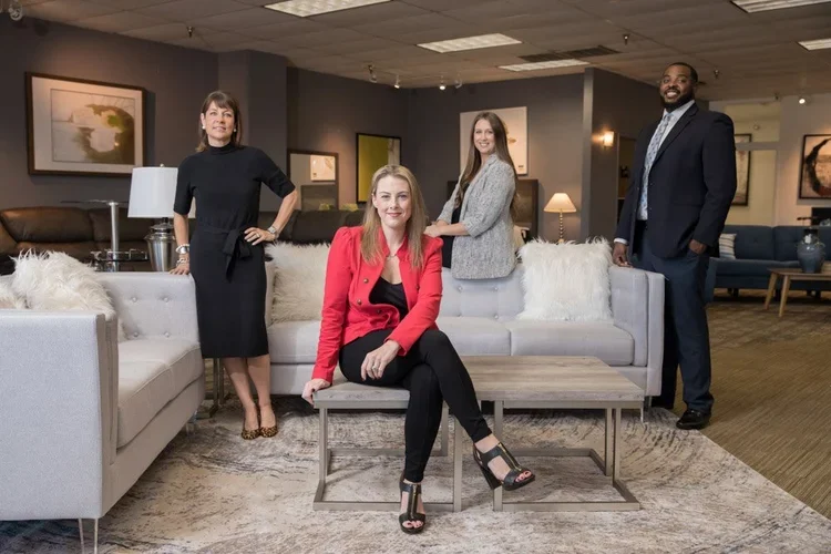 Four professionally dressed people posing in a modern living room with artwork, lamps, and a neutral color palette.