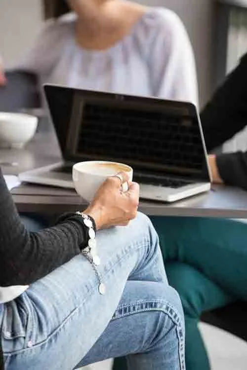 Person holding a coffee cup while sitting at a table with a laptop and other people.