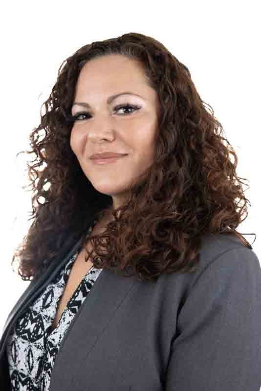 A woman with curly brown hair, wearing a gray blazer over a patterned blouse, smiling at the camera against a plain white background.
