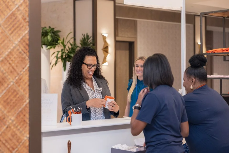 Four women at a store counter, one with glasses and curly hair holding an orange and white device, all smiling and engaged in conversation.