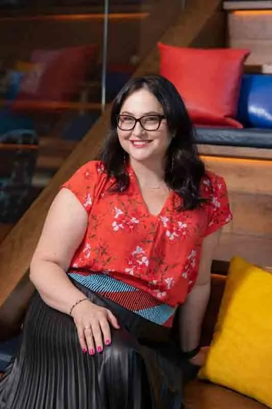 A smiling woman with glasses and dark hair, wearing a red floral blouse and striped skirt, sitting on a wooden staircase with colorful pillows in the background.