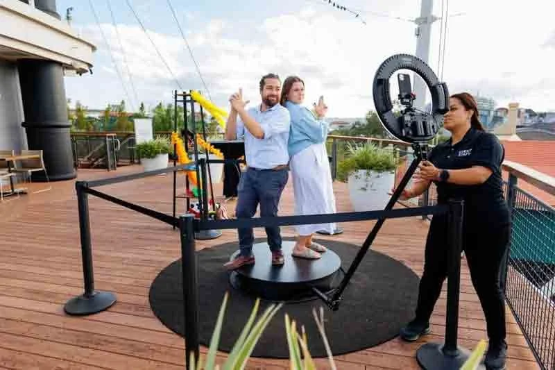 Two people pose on a small rotating platform on a rooftop with a professional videographer filming them with a ring light; the man is flipping a peace sign, and the woman is pointing with both hands, inside a designated filming area with black stanch