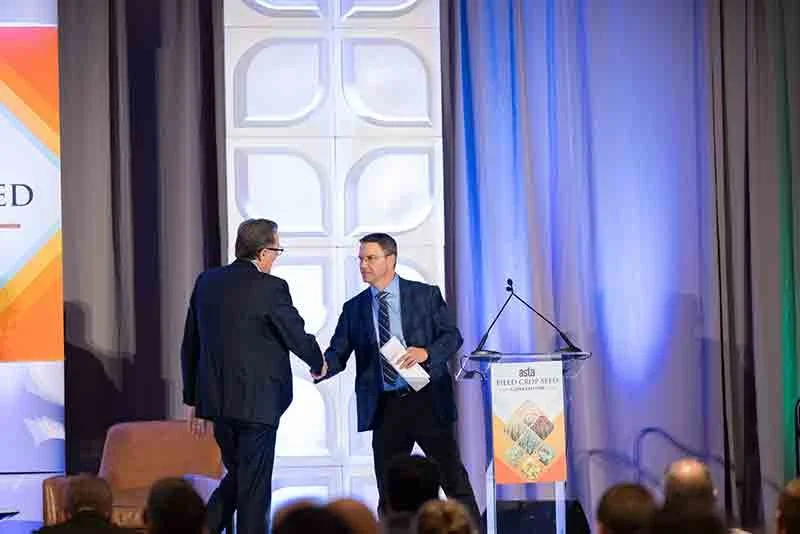 Two men in business suits shaking hands on a stage at a conference, with a podium and audience in front.