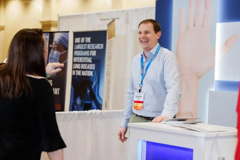 A man at a booth conversing with a woman at a conference or expo, with banners and informational signs in the background.