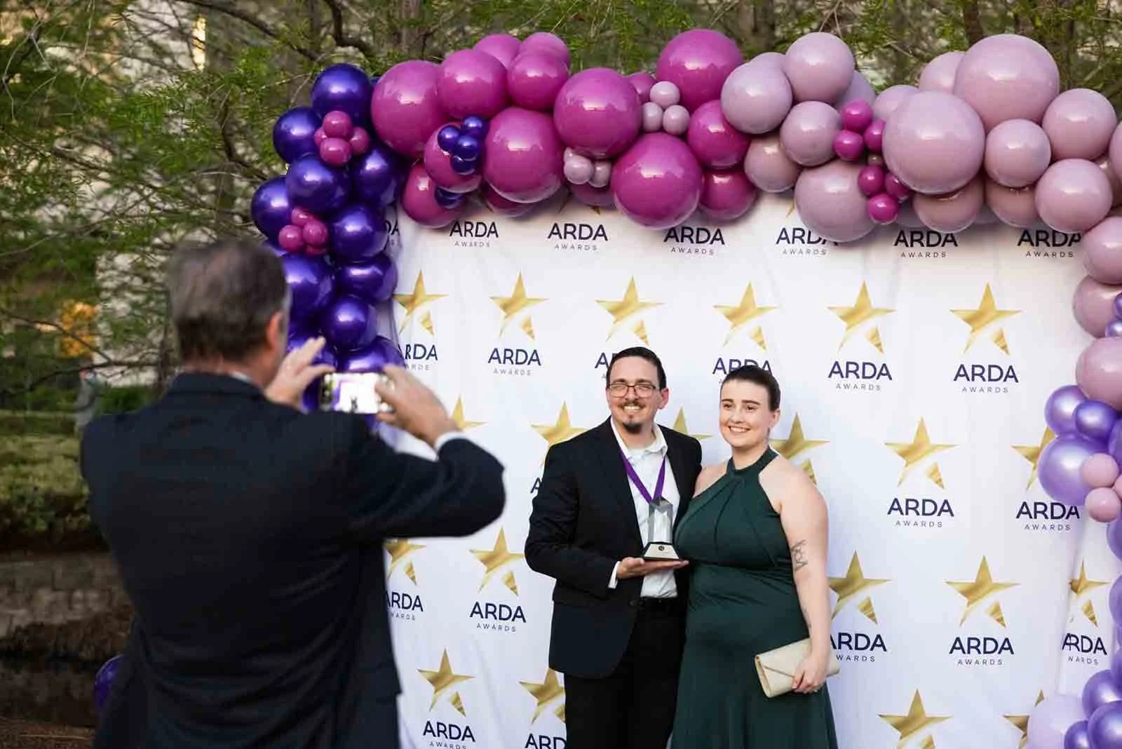 Two people in formal attire posing with an award at an ARDA Awards event, with a backdrop decorated with purple and pink balloons and the ARDA logo.