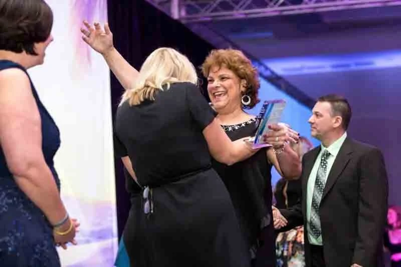 A group of people at a celebration or award event, with a woman receiving a trophy and smiling as others watch.