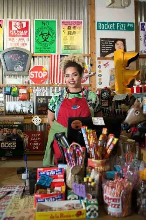 A woman behind a checkout counter in a store, surrounded by various gift items, toys, and posters on the wall, including a stop sign and a green biohazard sign. She is wearing a red apron and smiling.