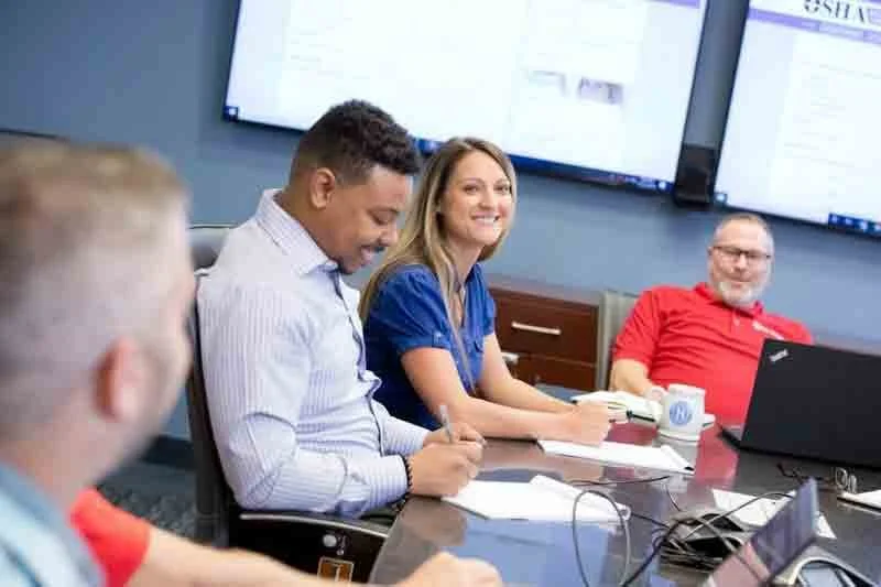 Group of four professionals in a meeting room, smiling and taking notes, with a large screen displaying a presentation behind them.