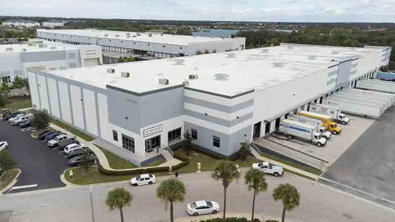 Large industrial warehouse with loading docks and trucks, surrounded by parking lot and palm trees.