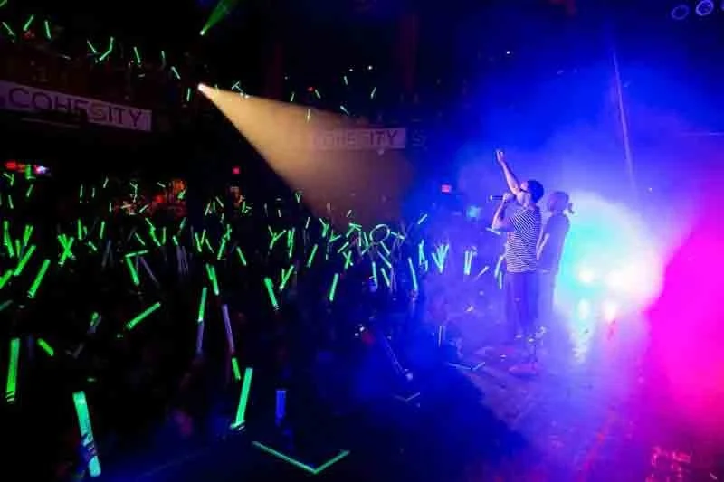 A concert scene with performers on stage and audience holding green glow sticks, colorful lighting and haze.