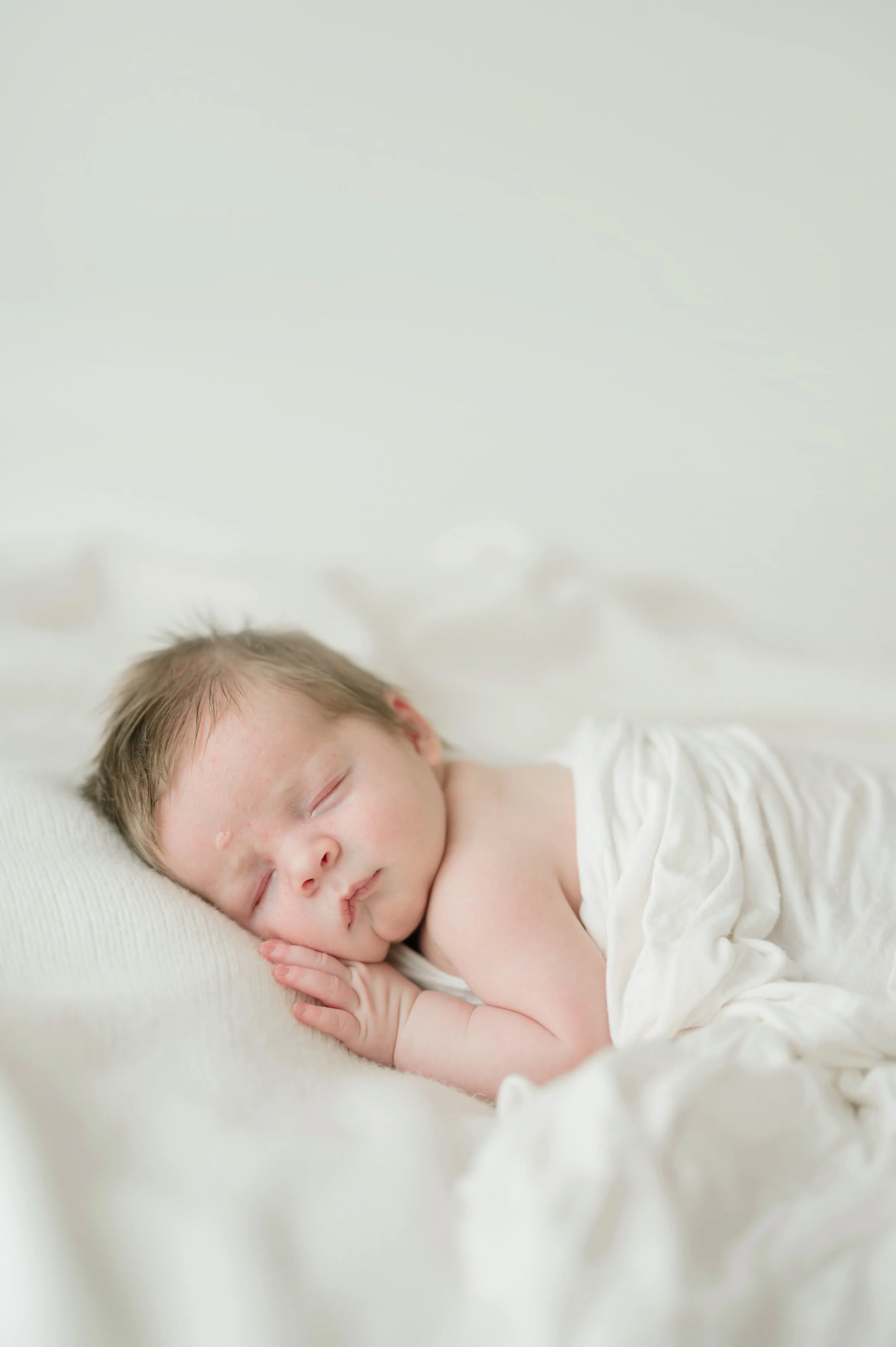 A young child with light skin and brown hair sleeping peacefully on a soft white surface, resting their head on their hands with a slight smile and wearing a white sleeveless top. Baton Rouge Maternity Newborn and Family Photographer 