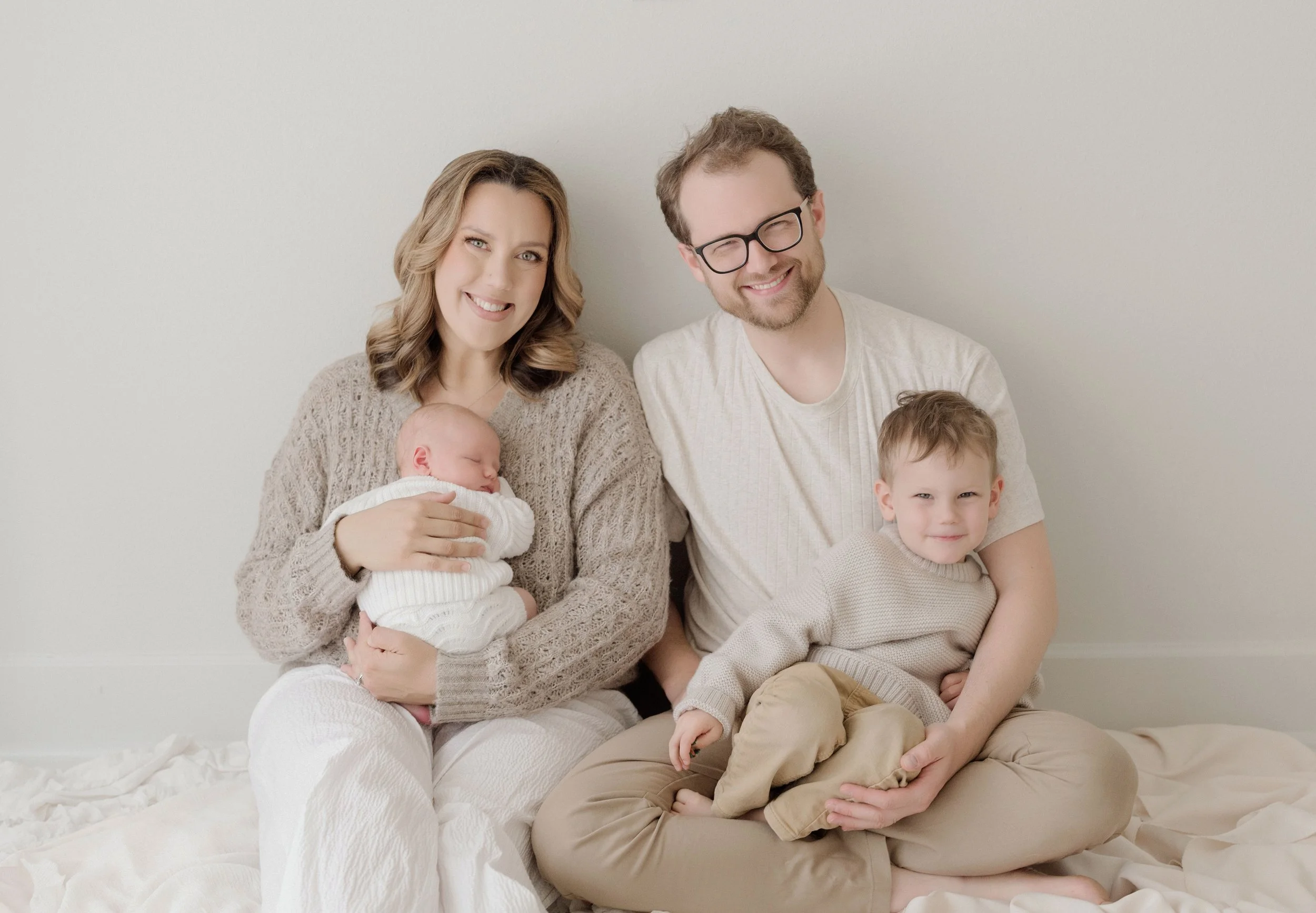 A family of four sitting on the floor against a plain white wall. The mother is holding a sleeping infant, and the father has two children, one sitting on his lap and another leaning against his shoulder. They are all smiling. Baton Rouge Maternity N