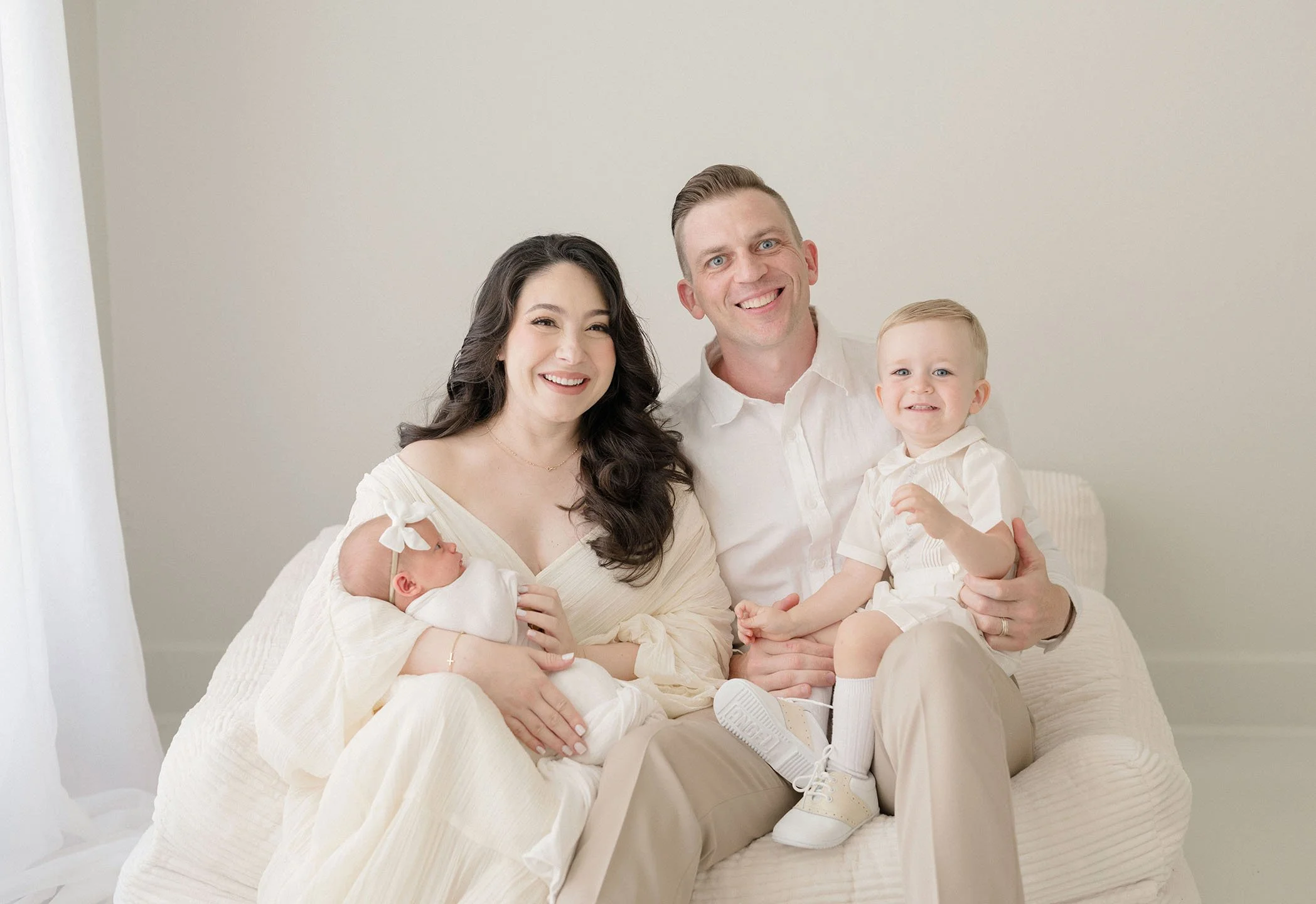 A happy family of four sitting on a white couch, with a woman holding a newborn and a man holding a young boy, all dressed in light-colored clothing against a neutral background.