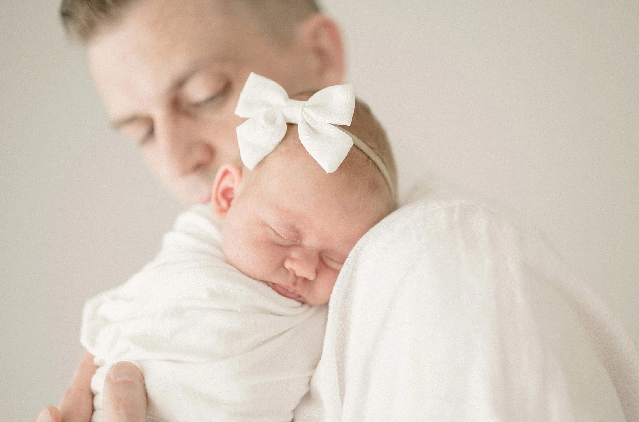 A person is holding a sleeping baby close, pressing their face gently against the baby's head, which is adorned with a white bow headband. The scene is soft and tender, with a neutral background.