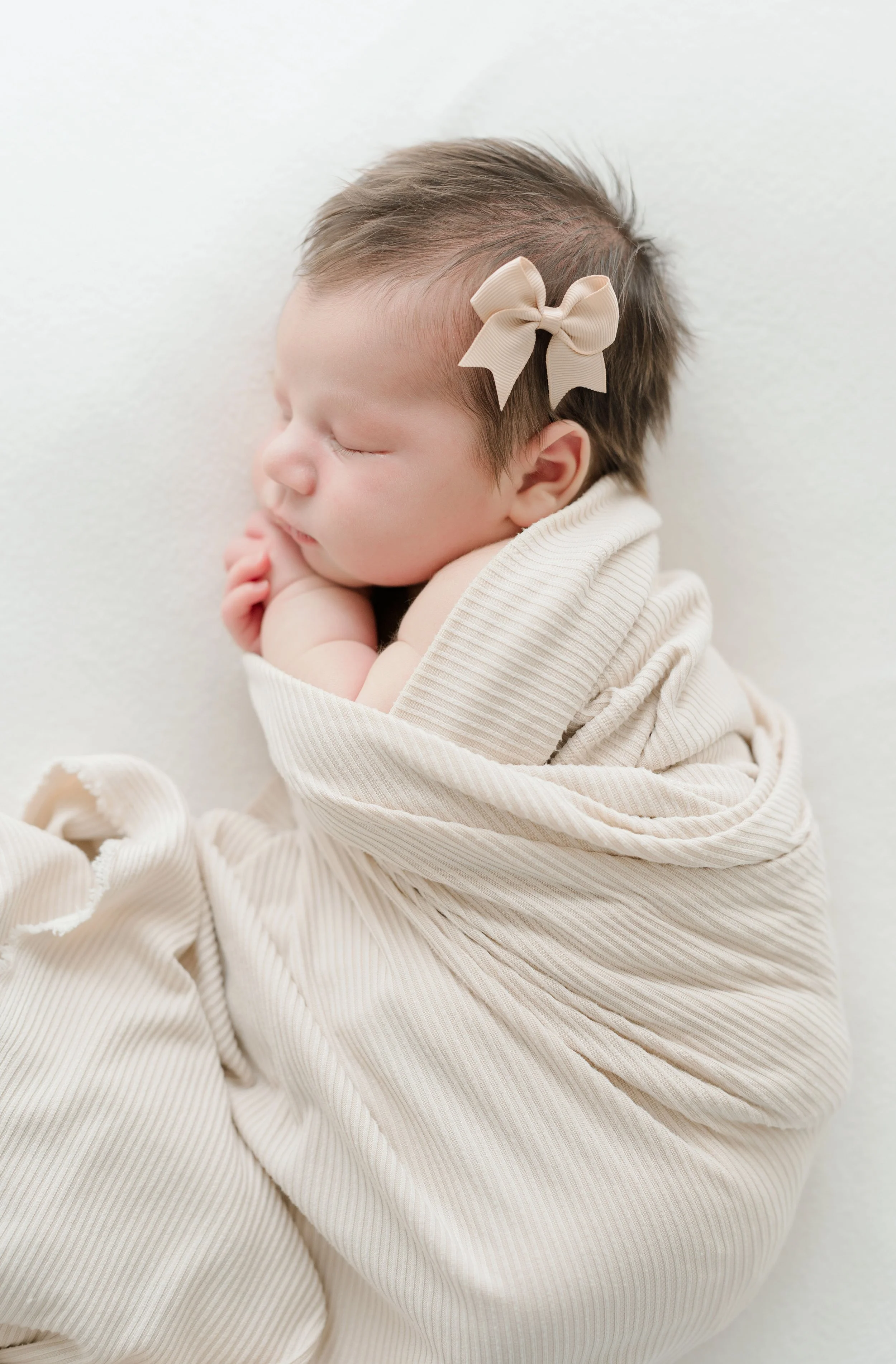 A sleeping baby with a beige bow in her hair, wrapped in a beige, ribbed blanket against a white background. Baton Rouge Maternity Newborn and Family Photographer 