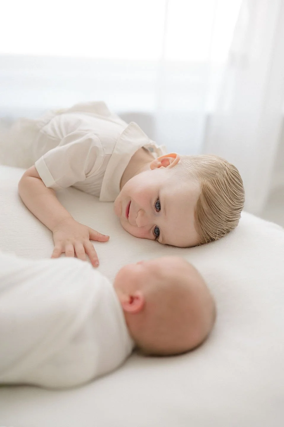 A young boy with blonde hair and blue eyes lying on a bed, looking at an infant with a shaved head, both in light-colored clothing.