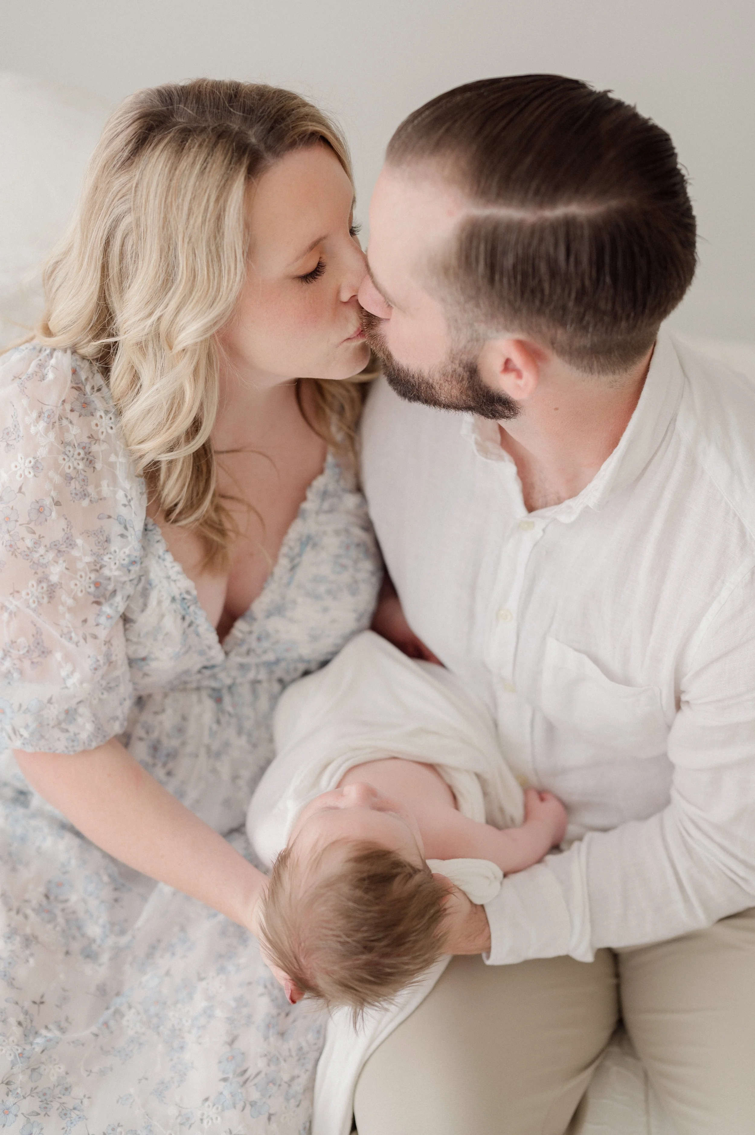 A family scene with two adults and a young child, all lying on a floral blanket, sharing a tender moment and touching noses and foreheads. Baton Rouge Maternity Newborn and Family Photographer 