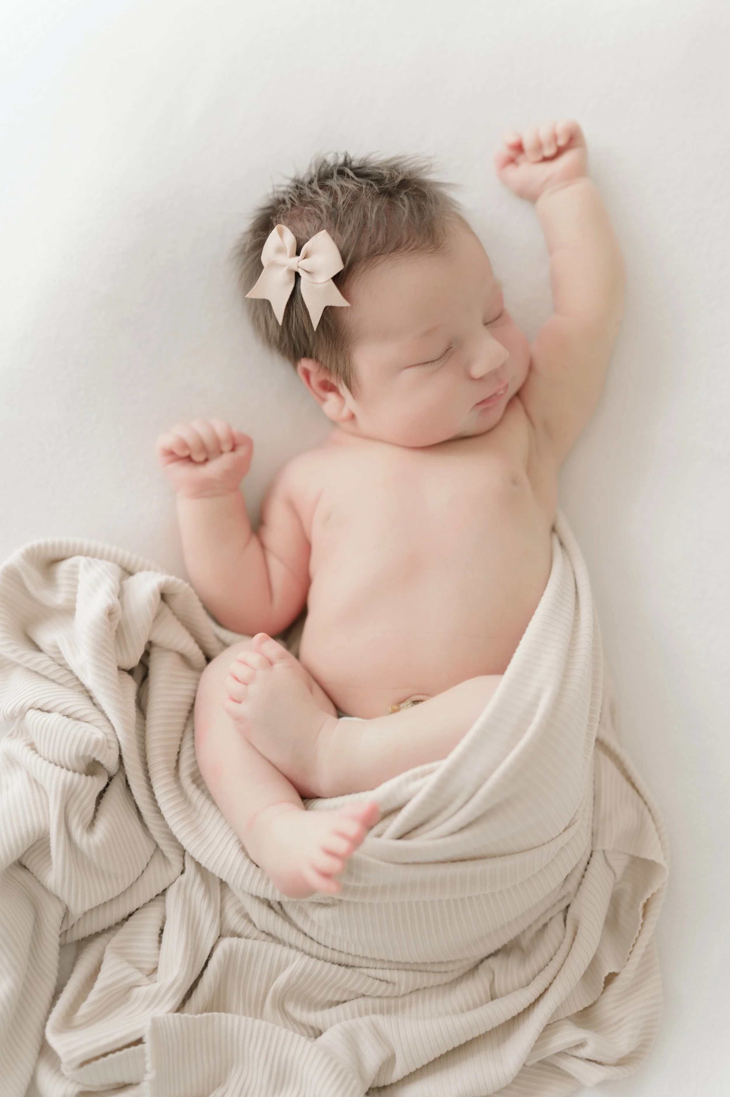A young baby with light brown hair, wearing a beige bow in her hair, is sleeping peacefully while lying on a soft white surface and wrapped in ribbed beige fabric. Baton Rouge Maternity Newborn and Family Photographer 