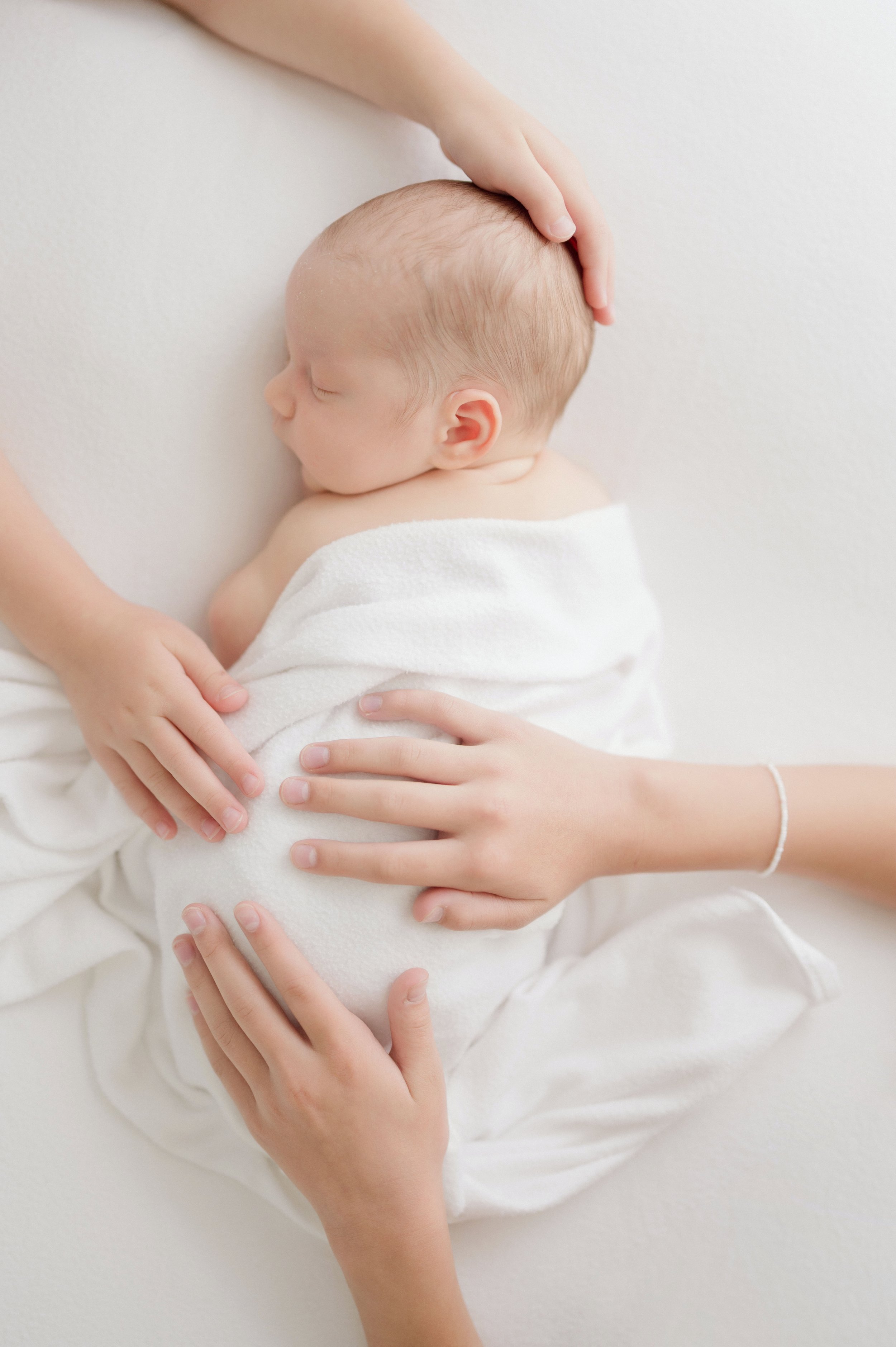 A person gently holding a sleeping baby wrapped in a white blanket, with a hand resting on the baby's head and another hand on its back, against a white background. Baton Rouge Maternity Newborn and Family Photographer 
