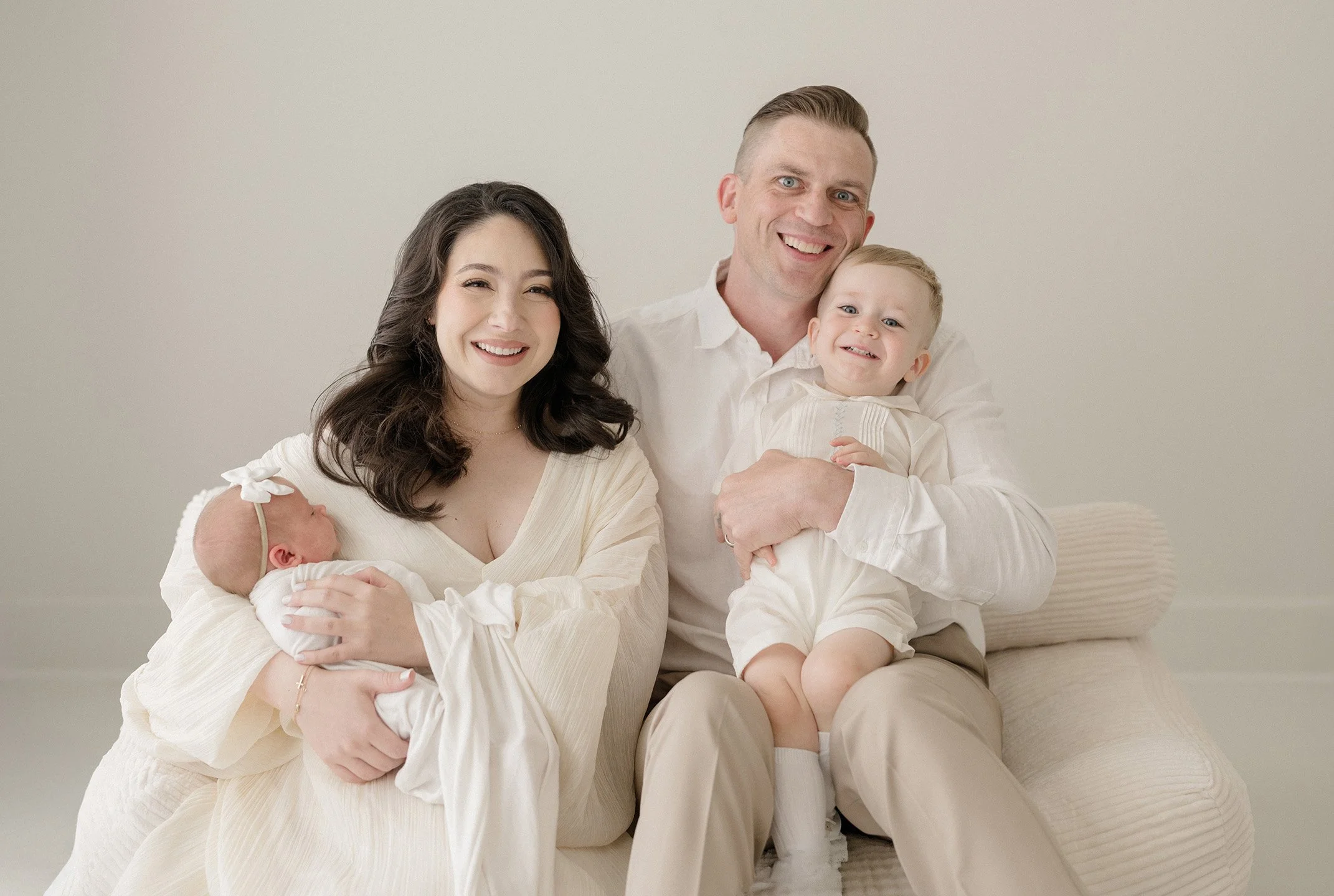 A happy family of four, including a mother, father, and two young children, sitting together in a neutral-colored room. The mother is holding a newborn, while the father holds an older child. All are smiling and dressed in light, coordinated clothing