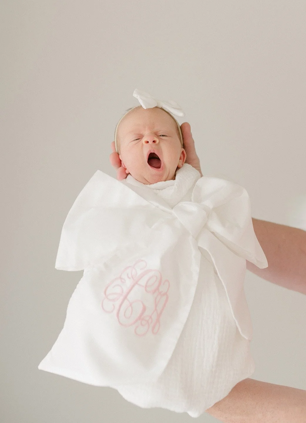 A newborn baby yawning while being held in a hand, wrapped in a white monogrammed blanket with a white bow headband.