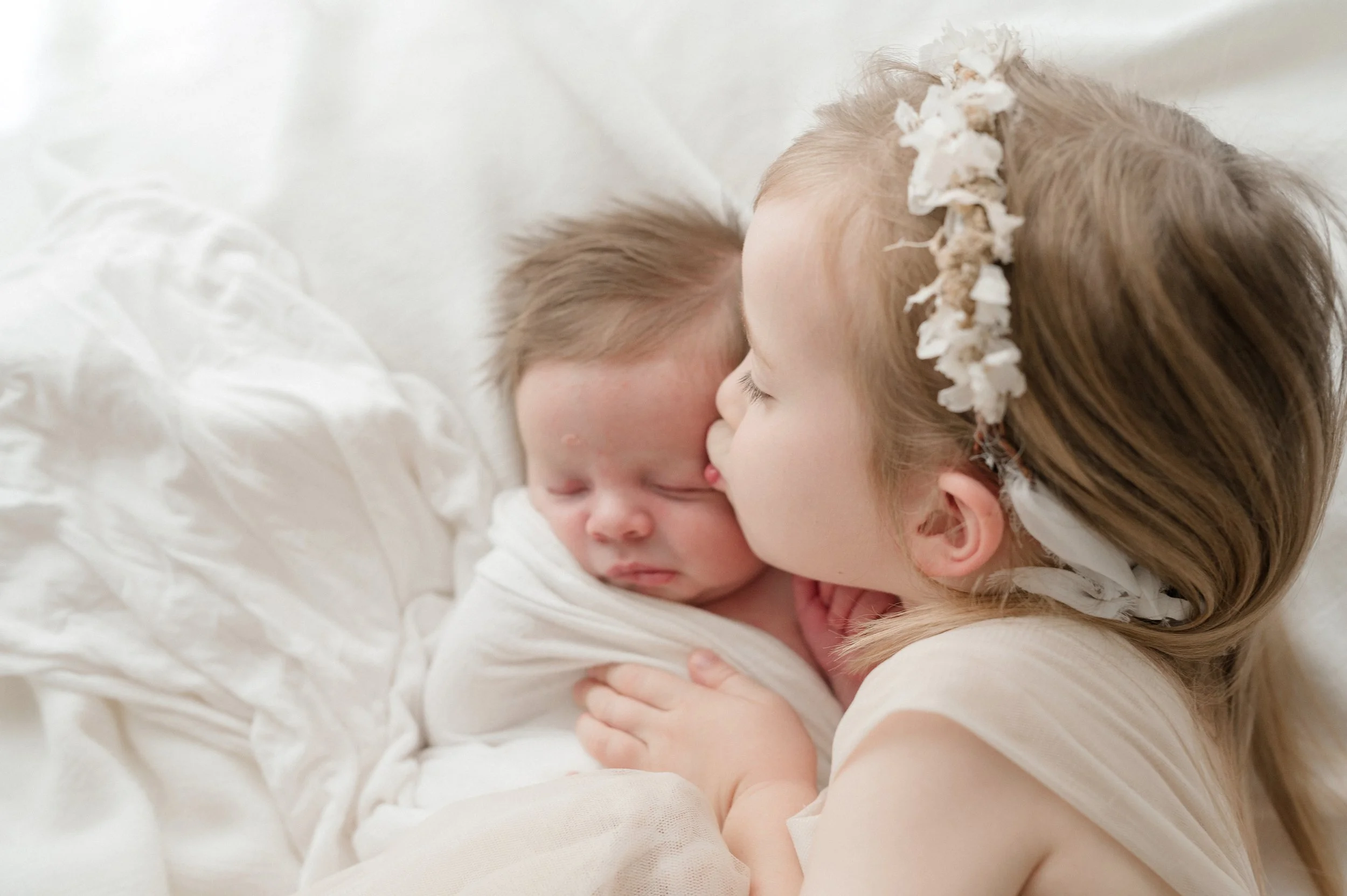 A young child with a floral headband kisses a sleeping baby on the forehead, both lying on a soft, white, textured blanket.