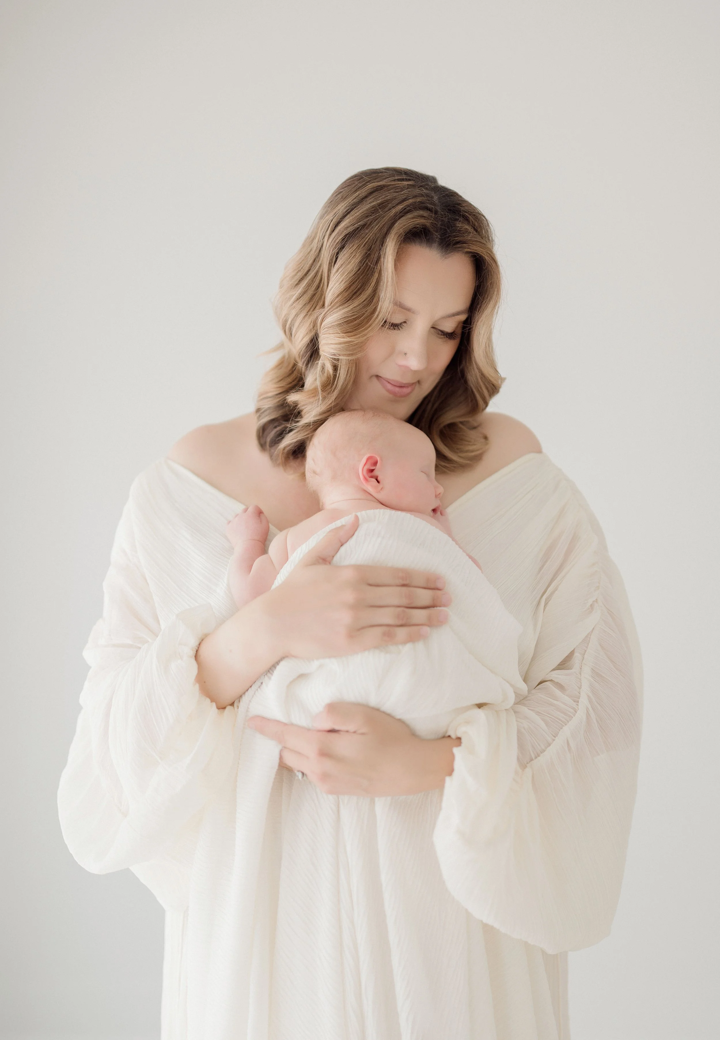 A woman with wavy light brown hair, dressed in a cream-colored flowing dress, holding and nuzzling a sleeping newborn baby wrapped in a matching white blanket, against a plain light background. Baton Rouge Maternity Newborn and Family Photographer 
