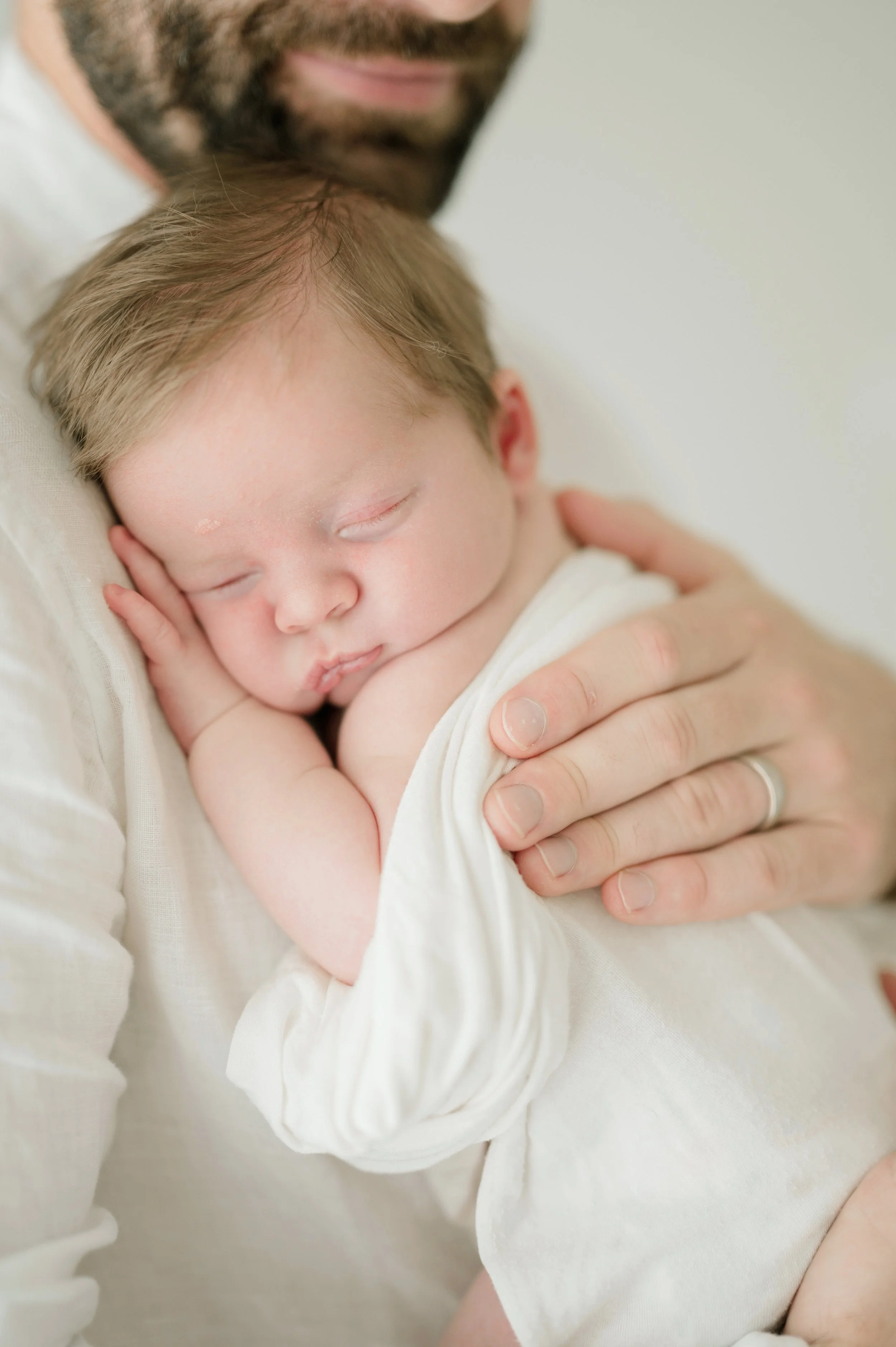 A close-up of a man holding a sleeping baby in his arms, with the baby's face resting on his hand. The baby has light hair and appears peaceful and content. Baton Rouge Maternity Newborn and Family Photographer 