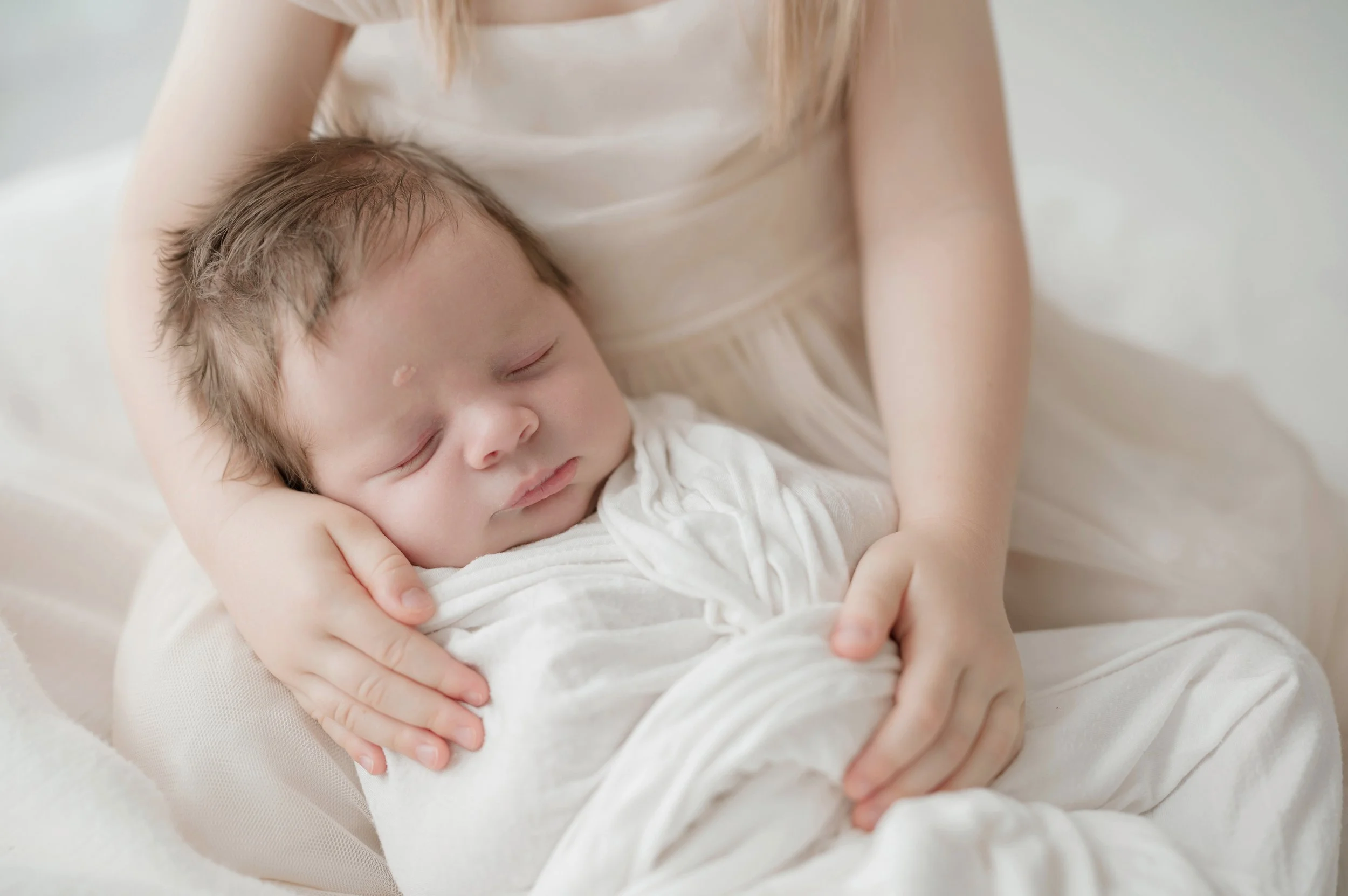 A young child with light brown hair sleeping peacefully, being held close by an adult with long blonde hair, wearing a cream-colored shirt. Baton Rouge Newborn Photographer 