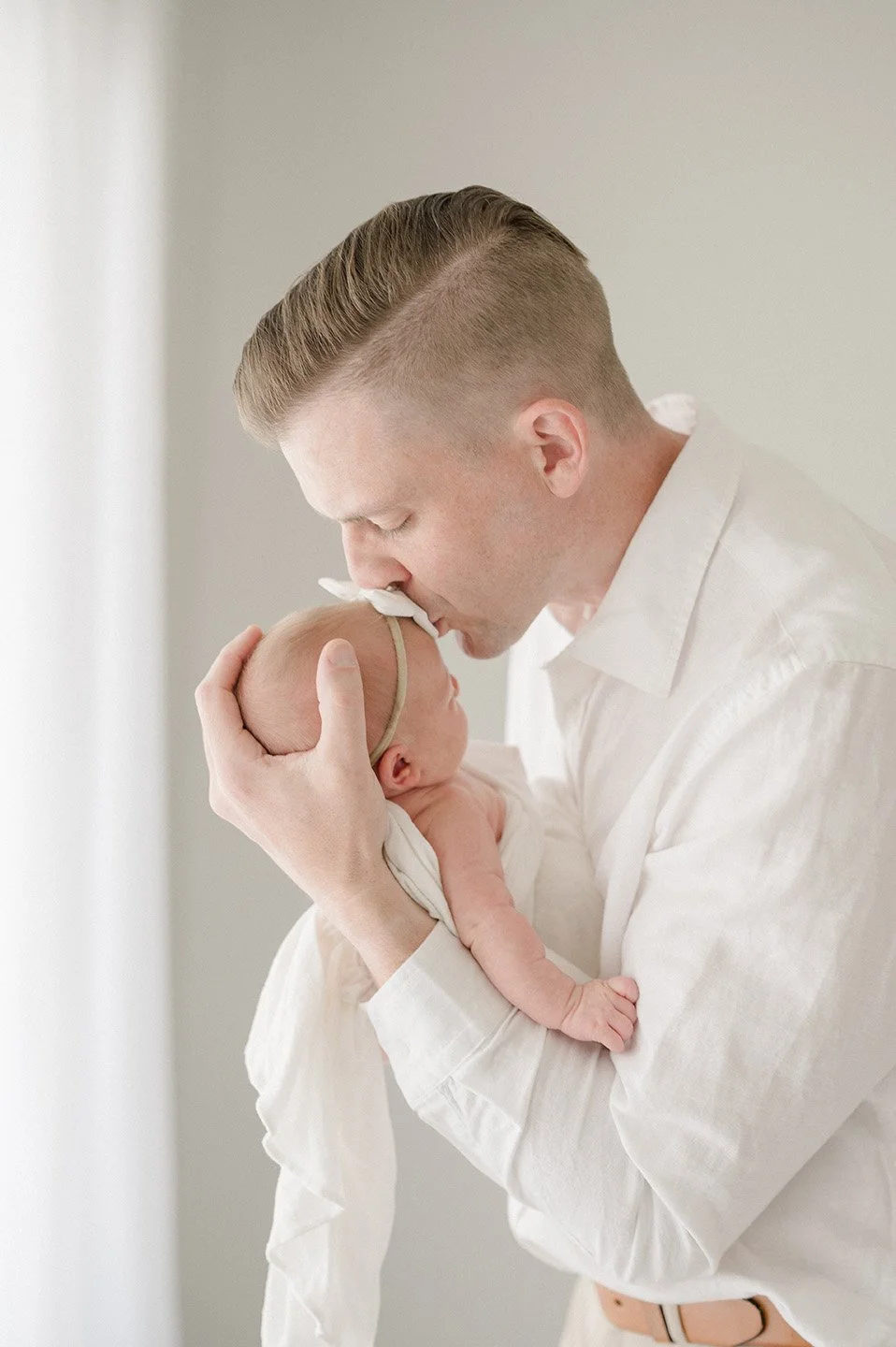 A man holding a newborn baby close, gently kissing the baby on the forehead in a softly lit room.