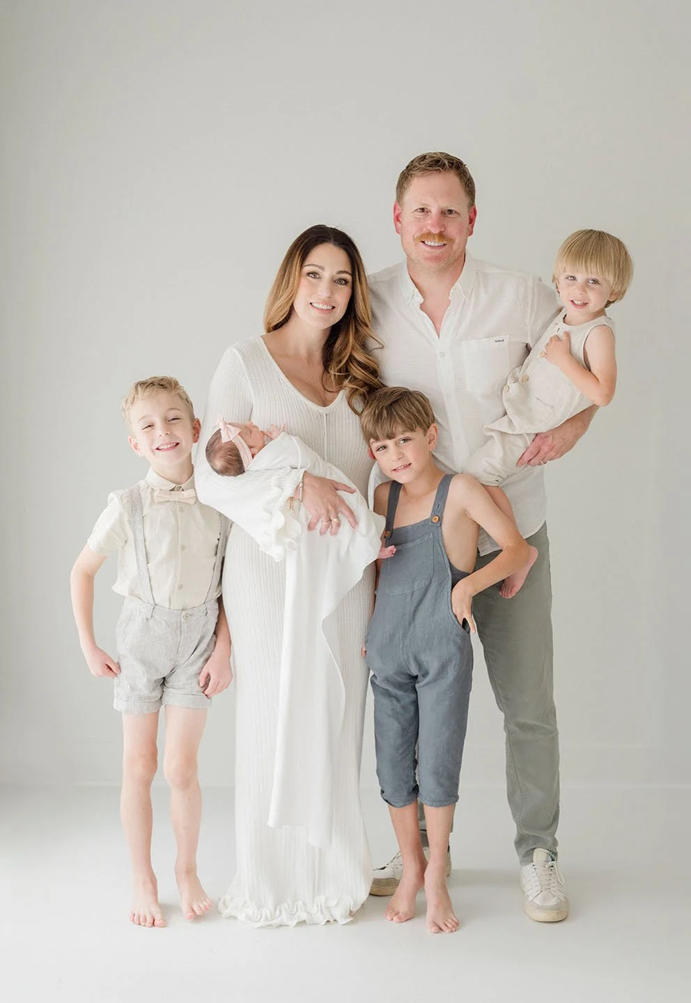 Family portrait of six people, including a mother, father, and four children, standing against a plain white background, with the mother holding a newborn baby. Baton Rouge Newborn and Family Photographer 
