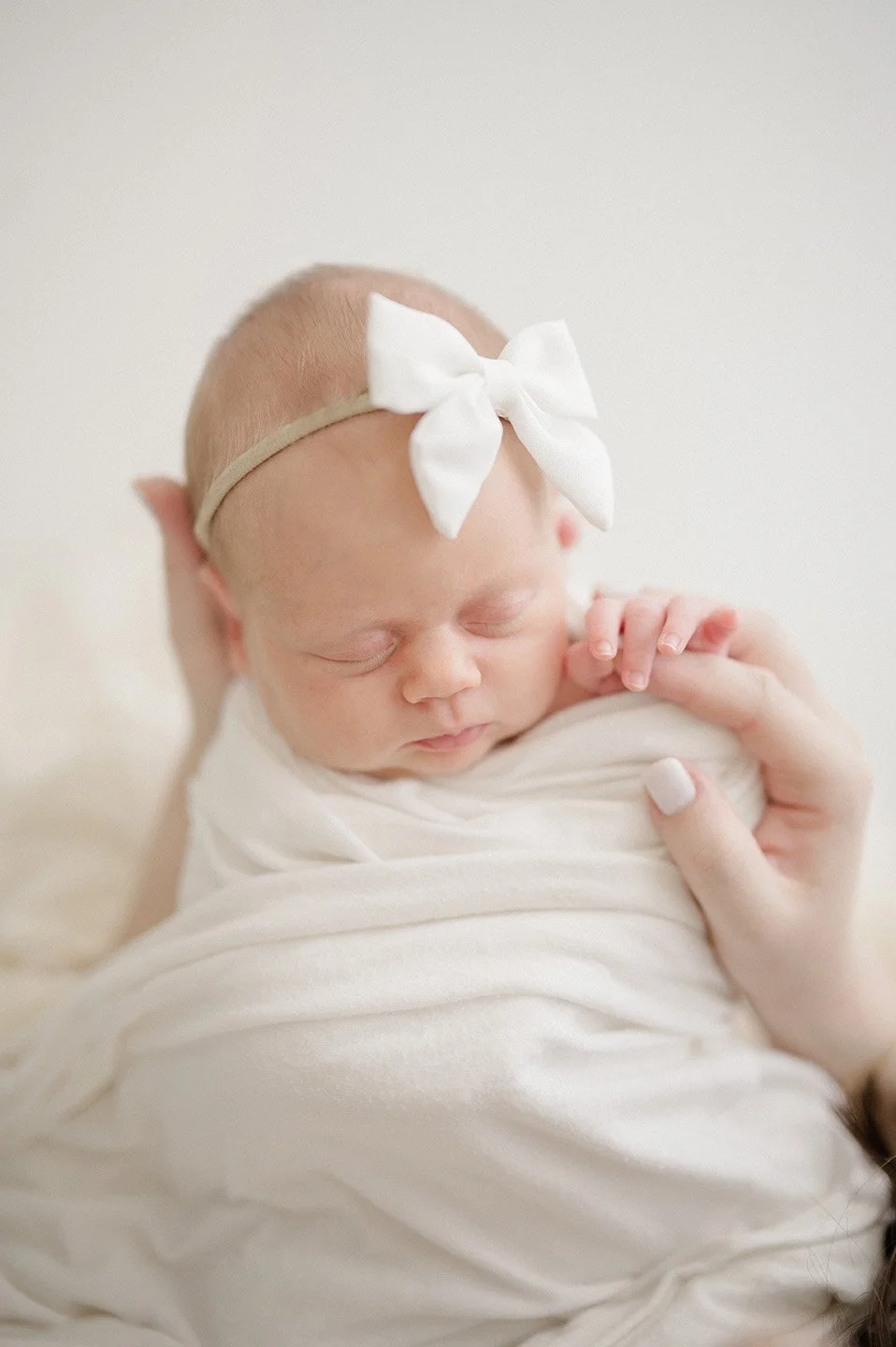 A sleeping newborn baby wearing a white headband with a big bow and wrapped in a white blanket. Baton Rouge Newborn Photographer 