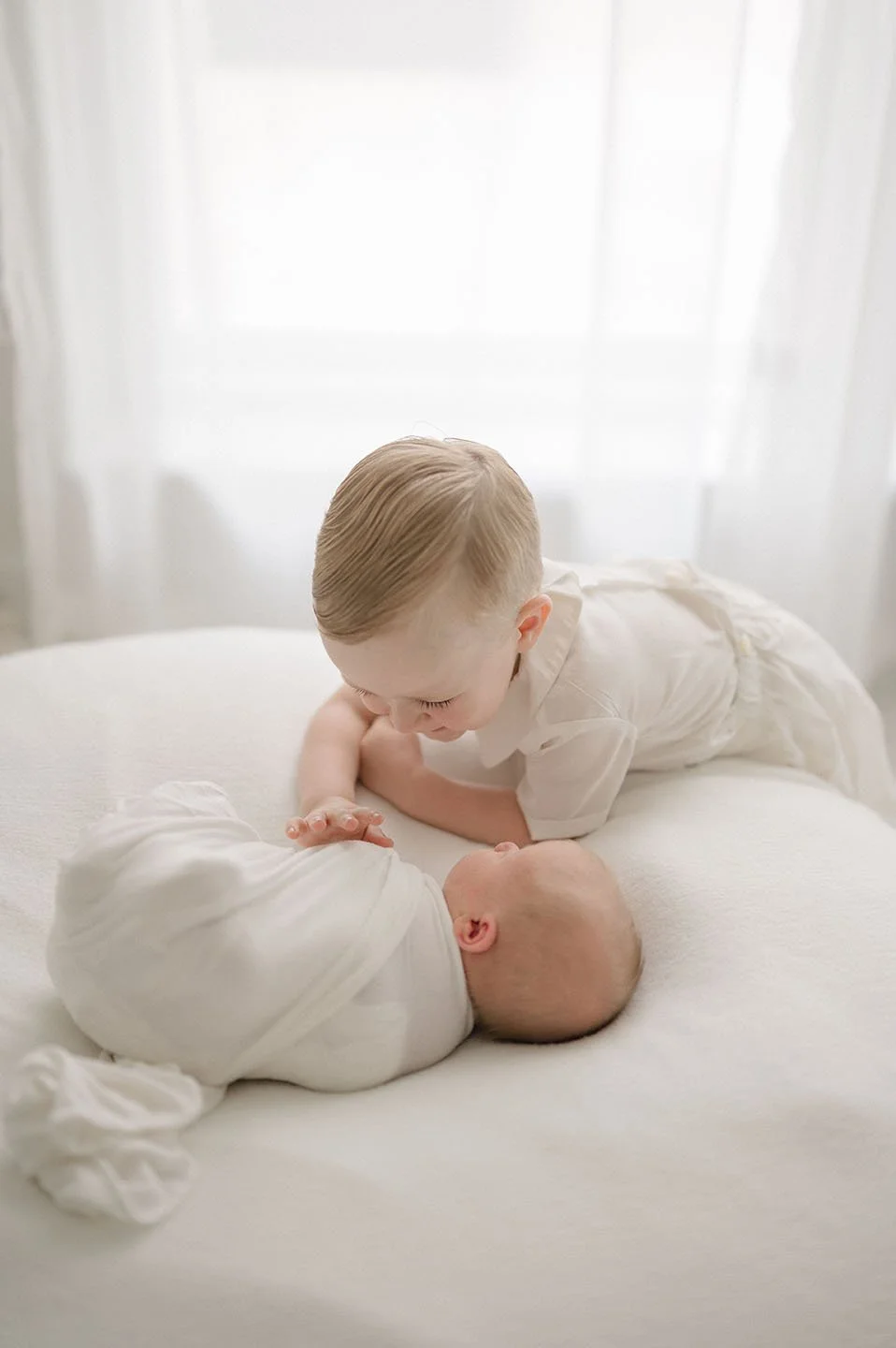 A young boy leaning over a bed, looking at a sleeping baby wrapped in a white blanket, in a bright room with white curtains.