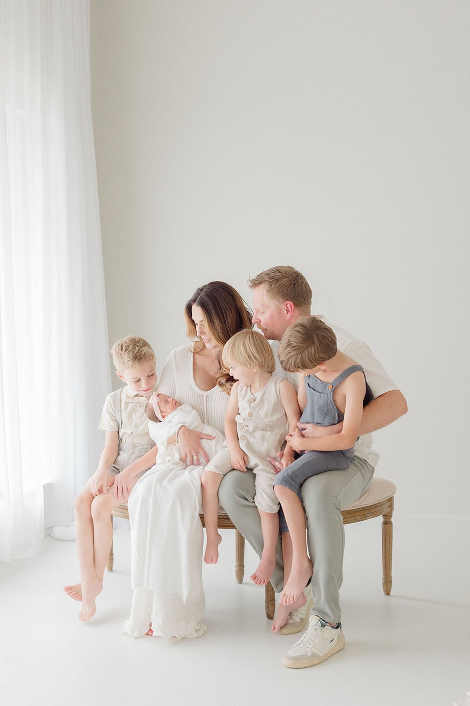 A family of six sitting on a bench in a bright, minimalistic room, with a woman holding a newborn and four young children gathered around her.  Baton Rouge Newborn Photographer Baton Rouge Newborn and Family Photographer 