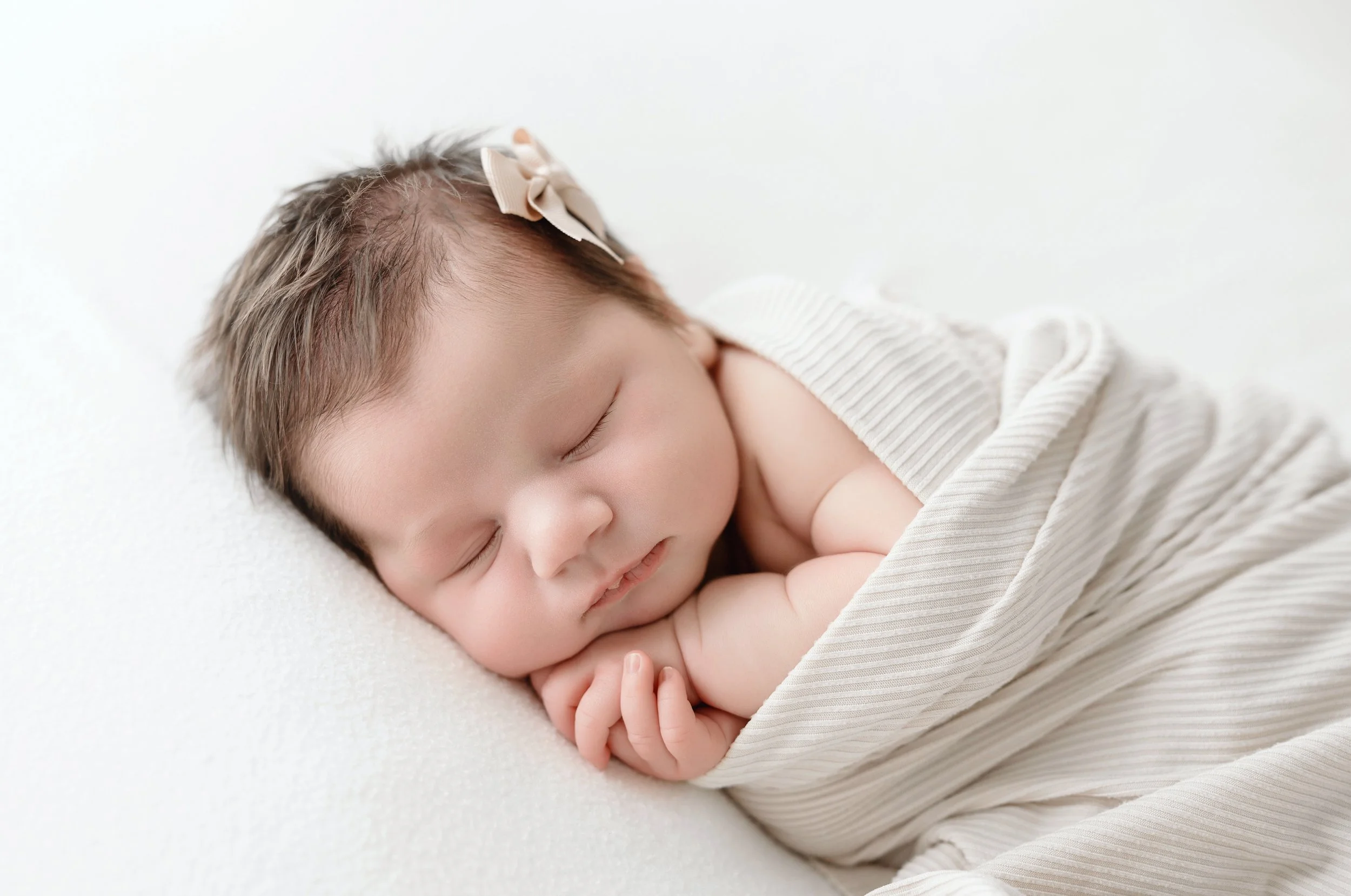 Close-up of a sleeping baby with light brown hair, wearing a beige bow, lying on a soft white surface, wrapped in a light beige striped blanket. Baton Rouge Maternity Newborn and Family Photographer 