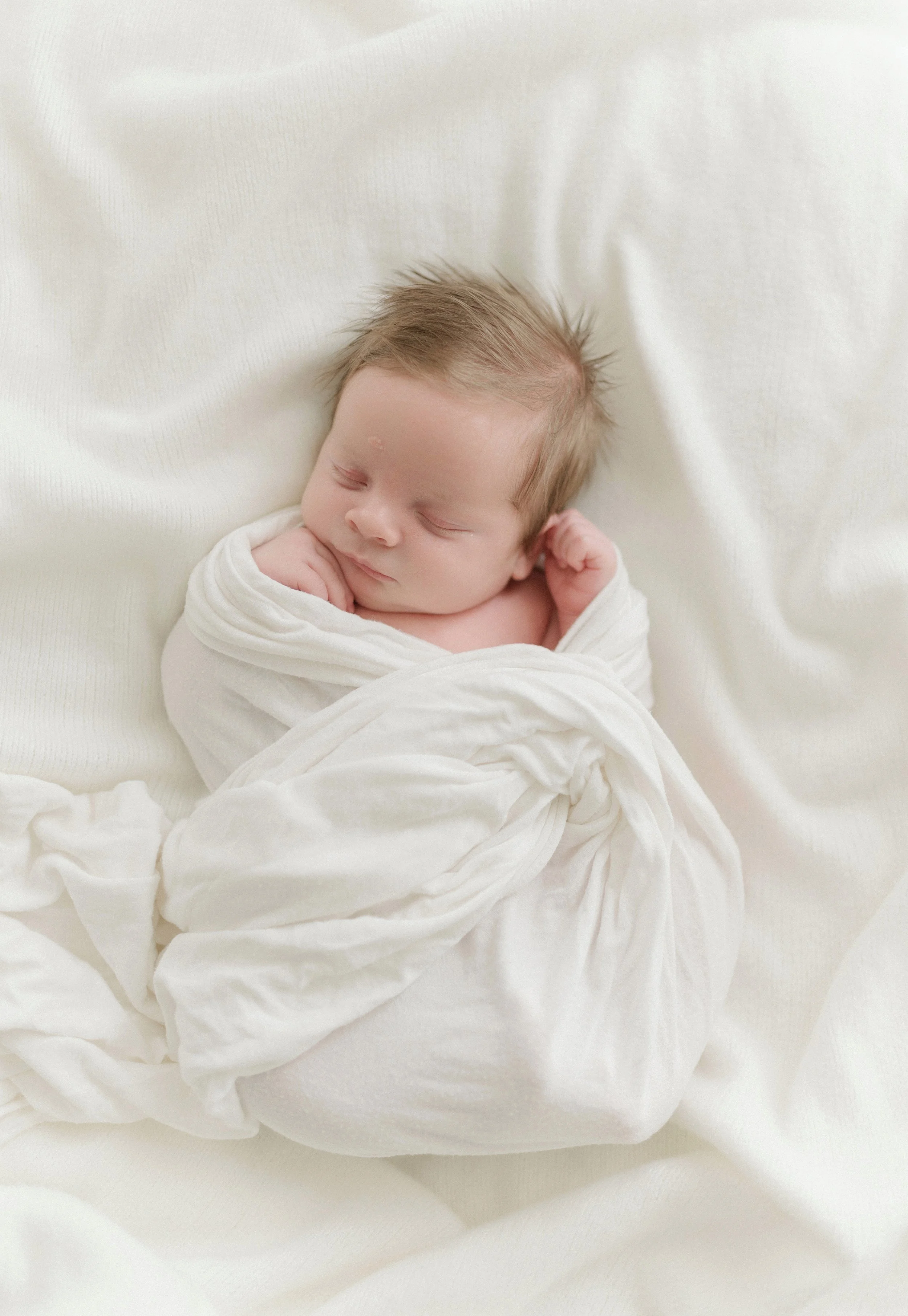 A newborn baby with closed eyes, wrapped in a white blanket, resting on a white surface. Baton Rouge Newborn Photographer 