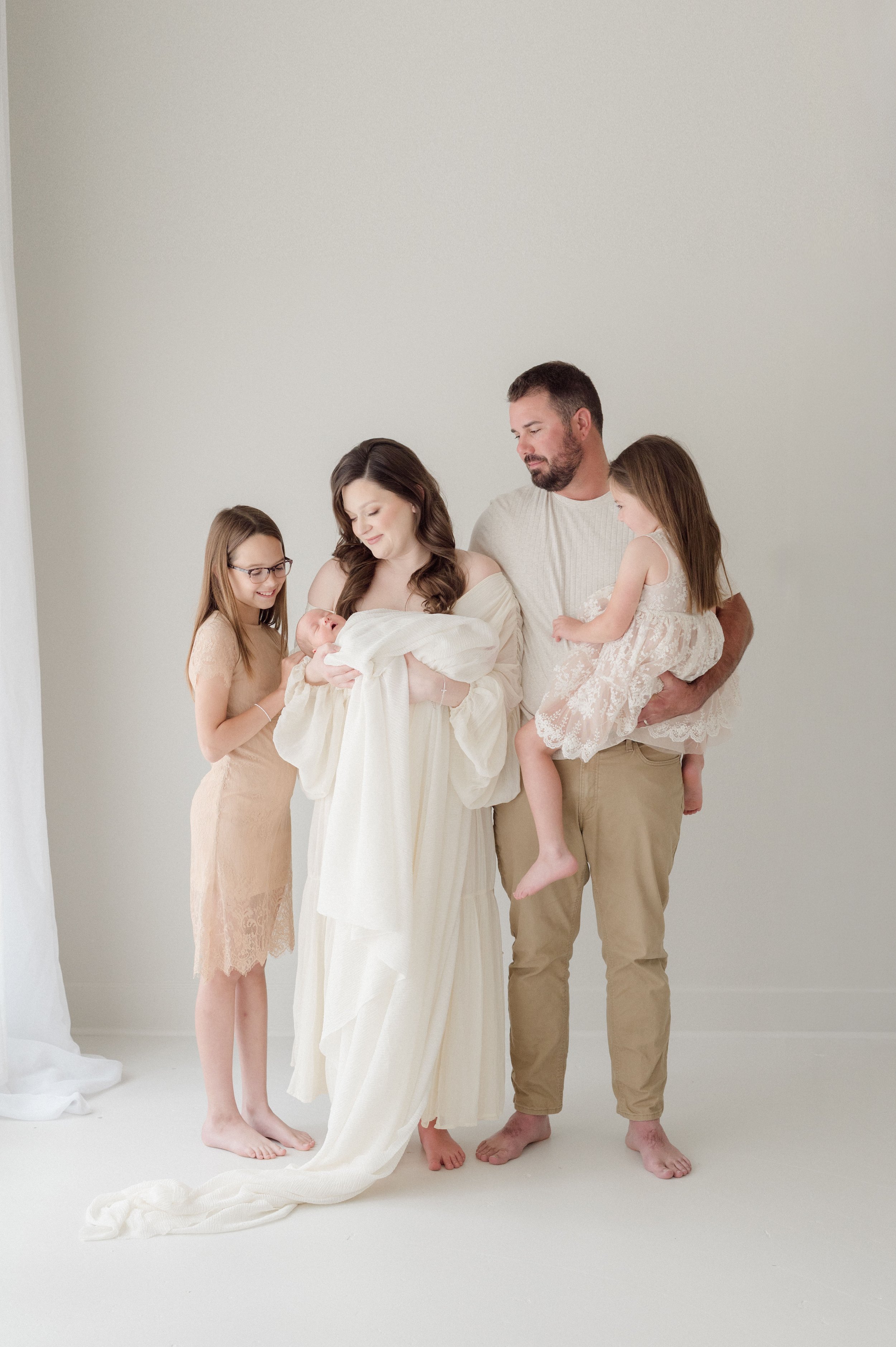 A family of five dressed in soft, neutral-colored clothing stands together against a plain white background Baton Rouge Maternity Newborn and Family Photographer 